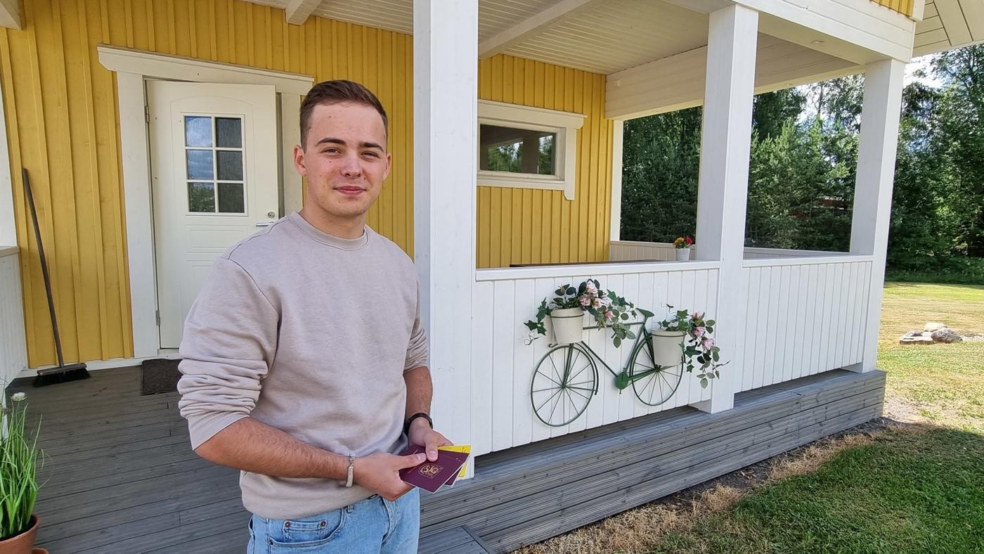 ung man med sjömanspass i handen vid gult trähus
