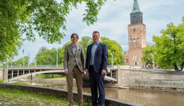 Två män iklädda kostym står vid Aura å, i bakgrunden syns Domkyrkan.