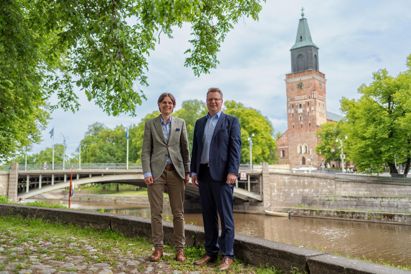 Två män iklädda kostym står vid Aura å, i bakgrunden syns Domkyrkan.