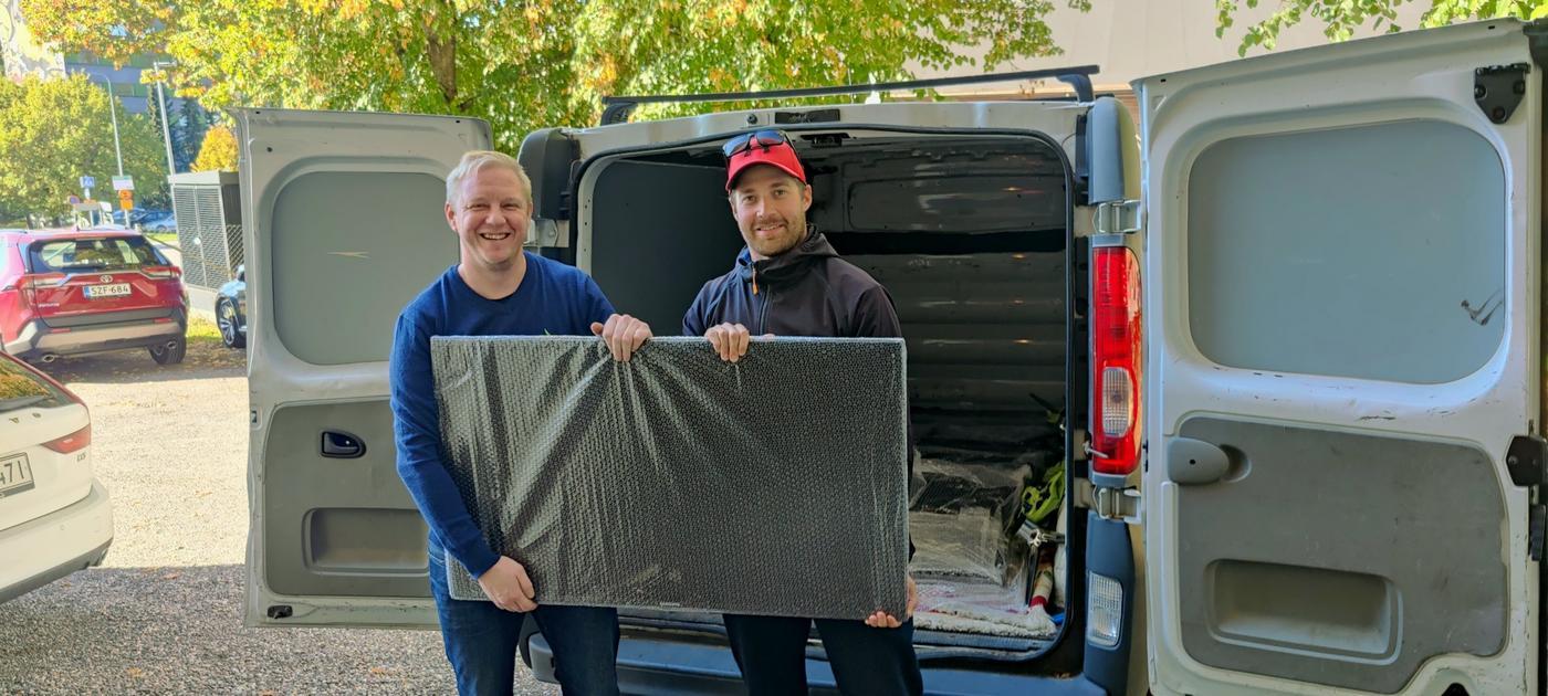 Två leende män håller upp en inpaketerad tv framför en paketbil.
