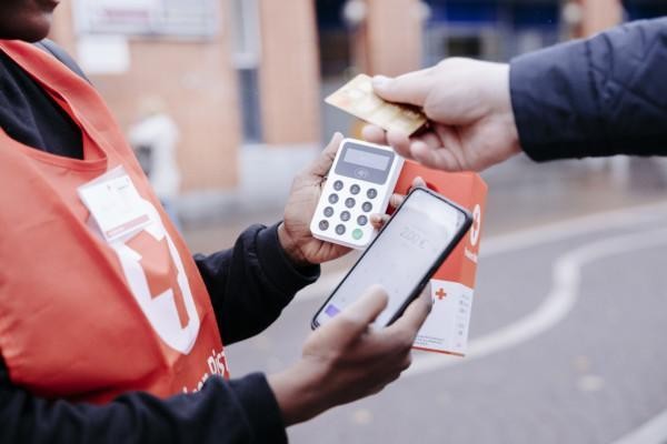 Person som håller i en insamlingsbössa och betalterminal, en annan persons hand sträcker sig från höger sida med ett bankkort i handen.