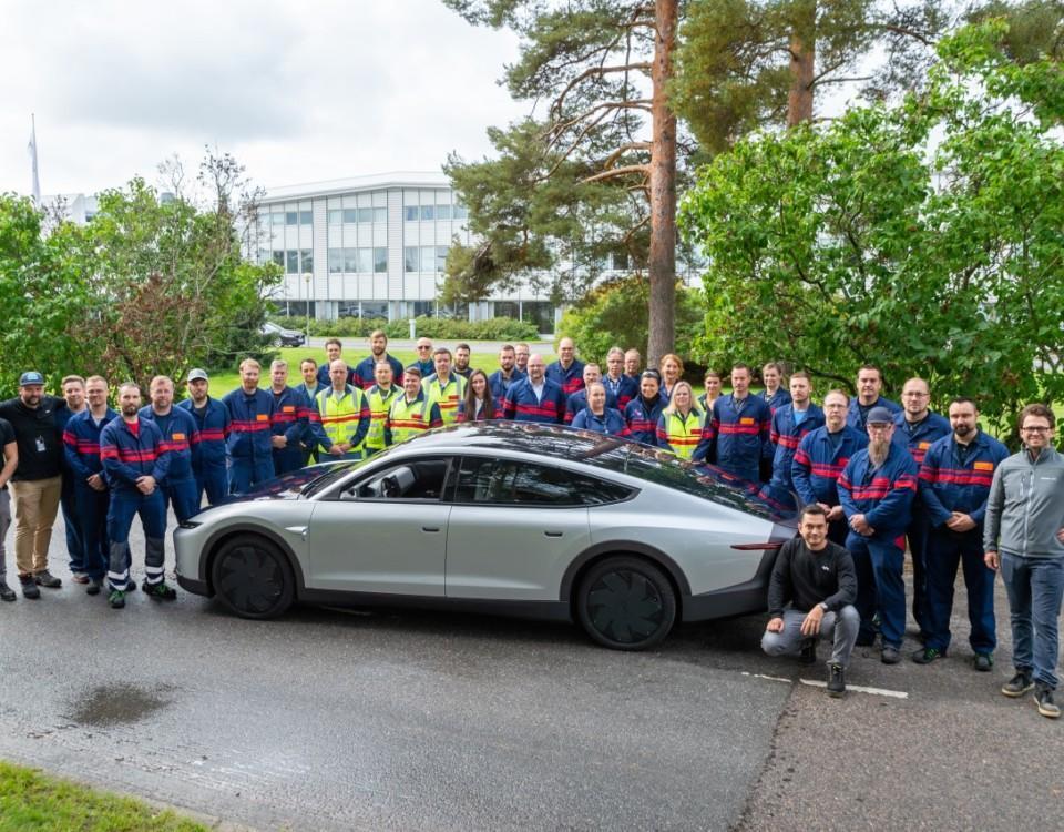Ungefär 40 personer står samlade kring en silverfärgad bil.