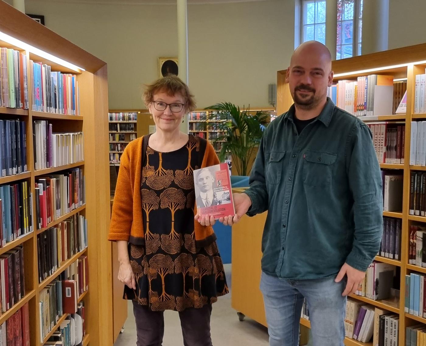 kvinna och man mellan bibliotekshyllor håller upp bok