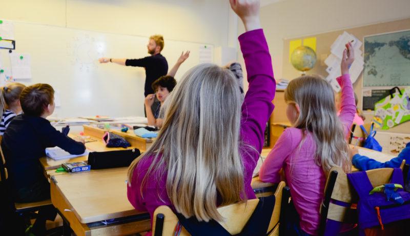 Flicka räcker upp handen och en lärare står vid tavlan.
