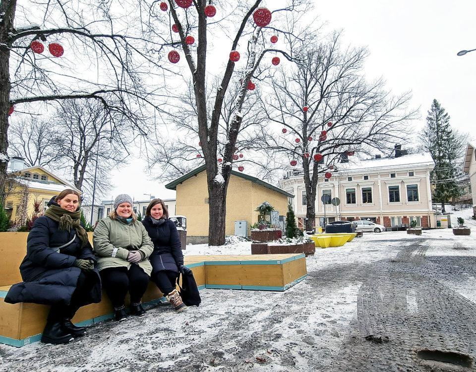Tre kvinnor på en bänk under träd pyntade med stora röda bollar.