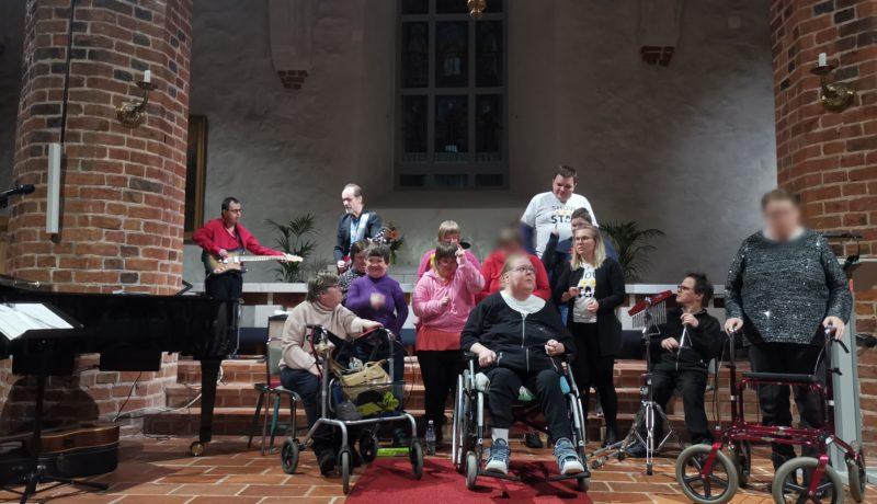 Ett gäng uppträder, vissa vid rollator/rullstol, framme i en kyrka.