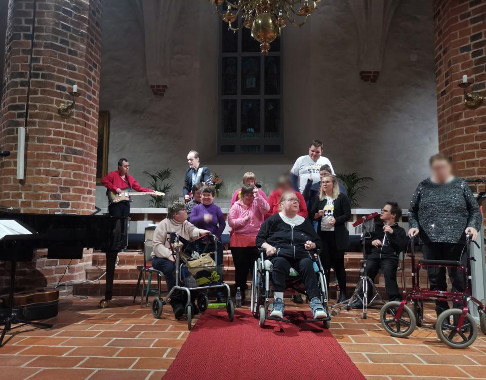 Ett gäng uppträder, vissa vid rollator/rullstol, framme i en kyrka.
