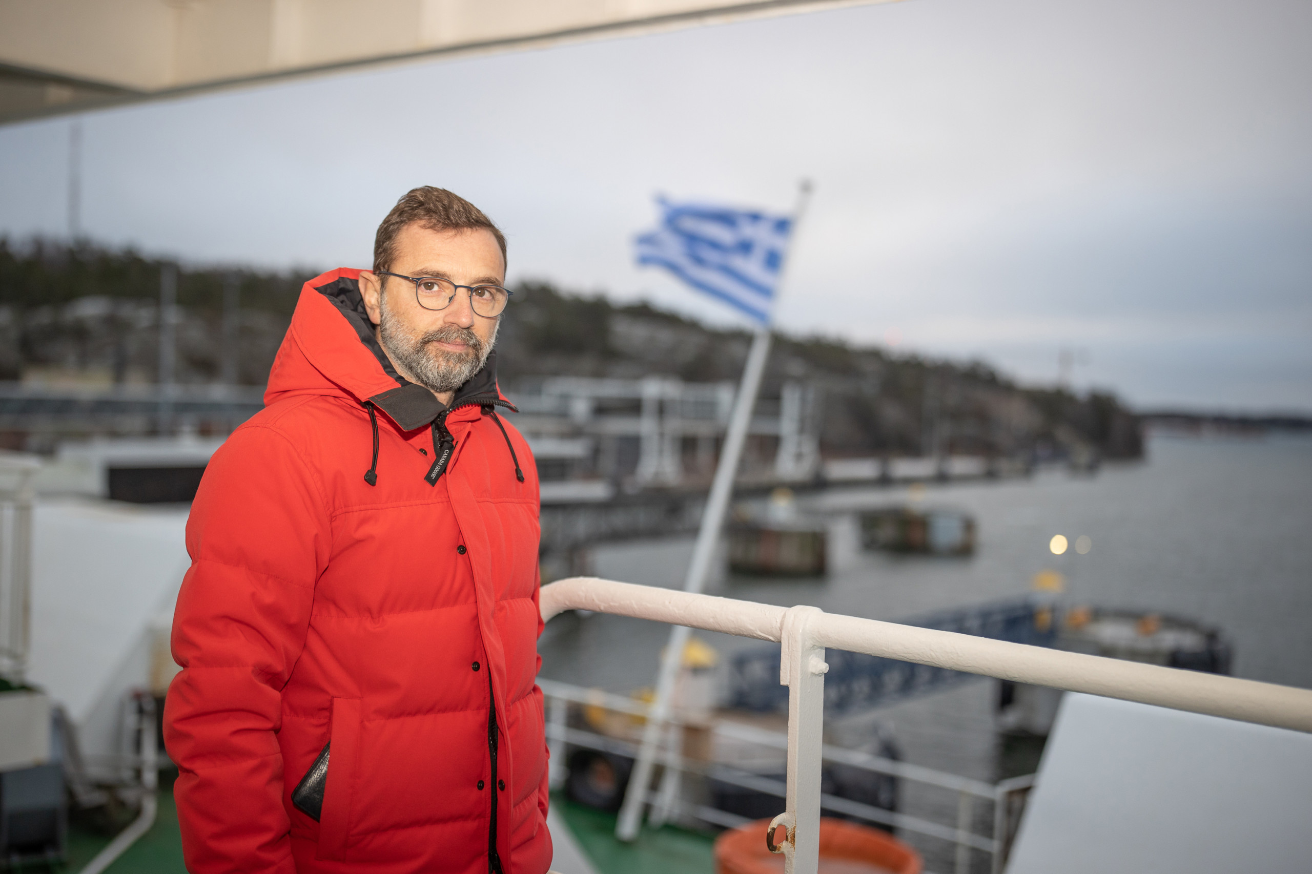 En man i röd täckjacka ombord på ett fartyg, i bakgrunden vajar Greklands flagga.