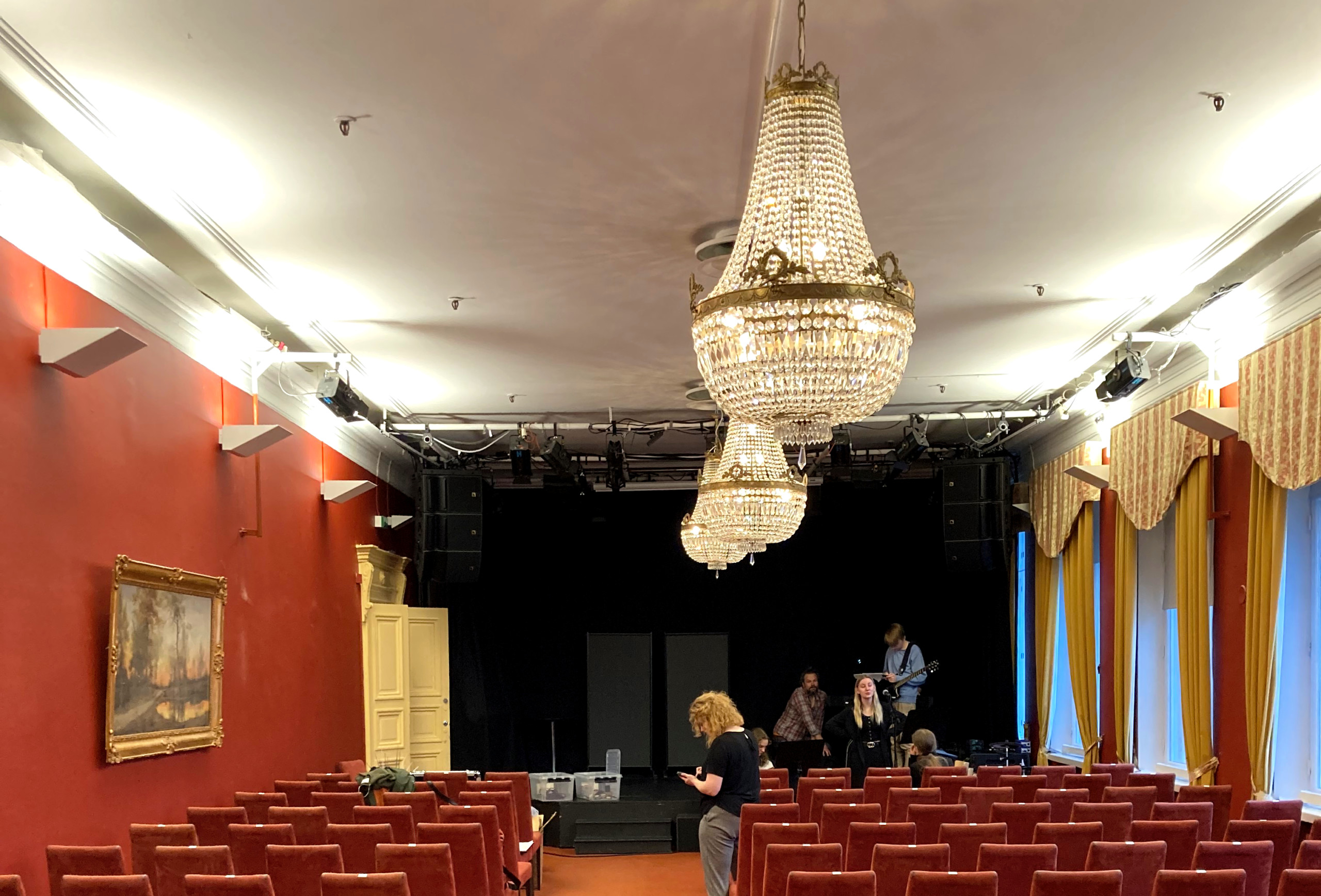 Teatersal med röda väggar, röda stolar och två stora kristallkronor i taket. Scenen längst bak har svart vägg.