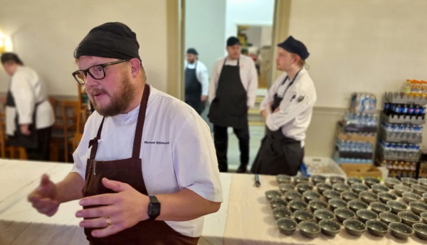 Kock gestikulerar med kärl och andra kockar bakgrunden.