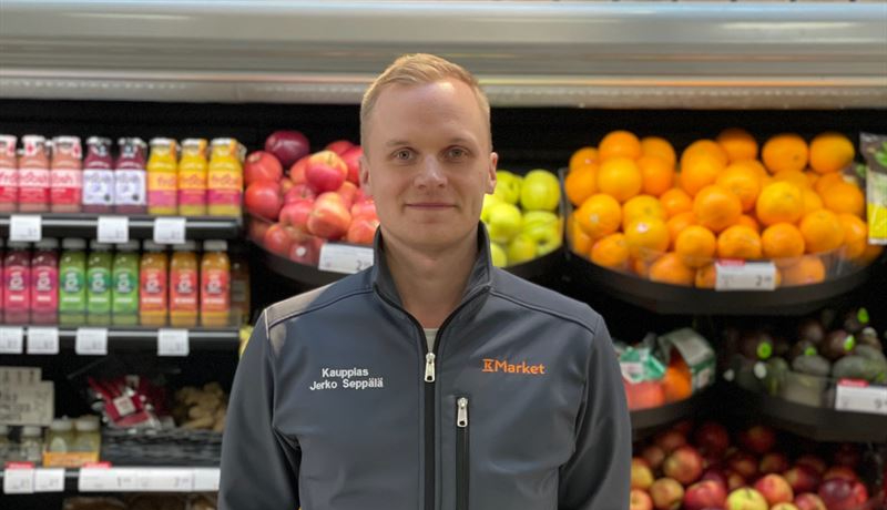 Man i grå jacka framför butikshylla med frukt och grön
