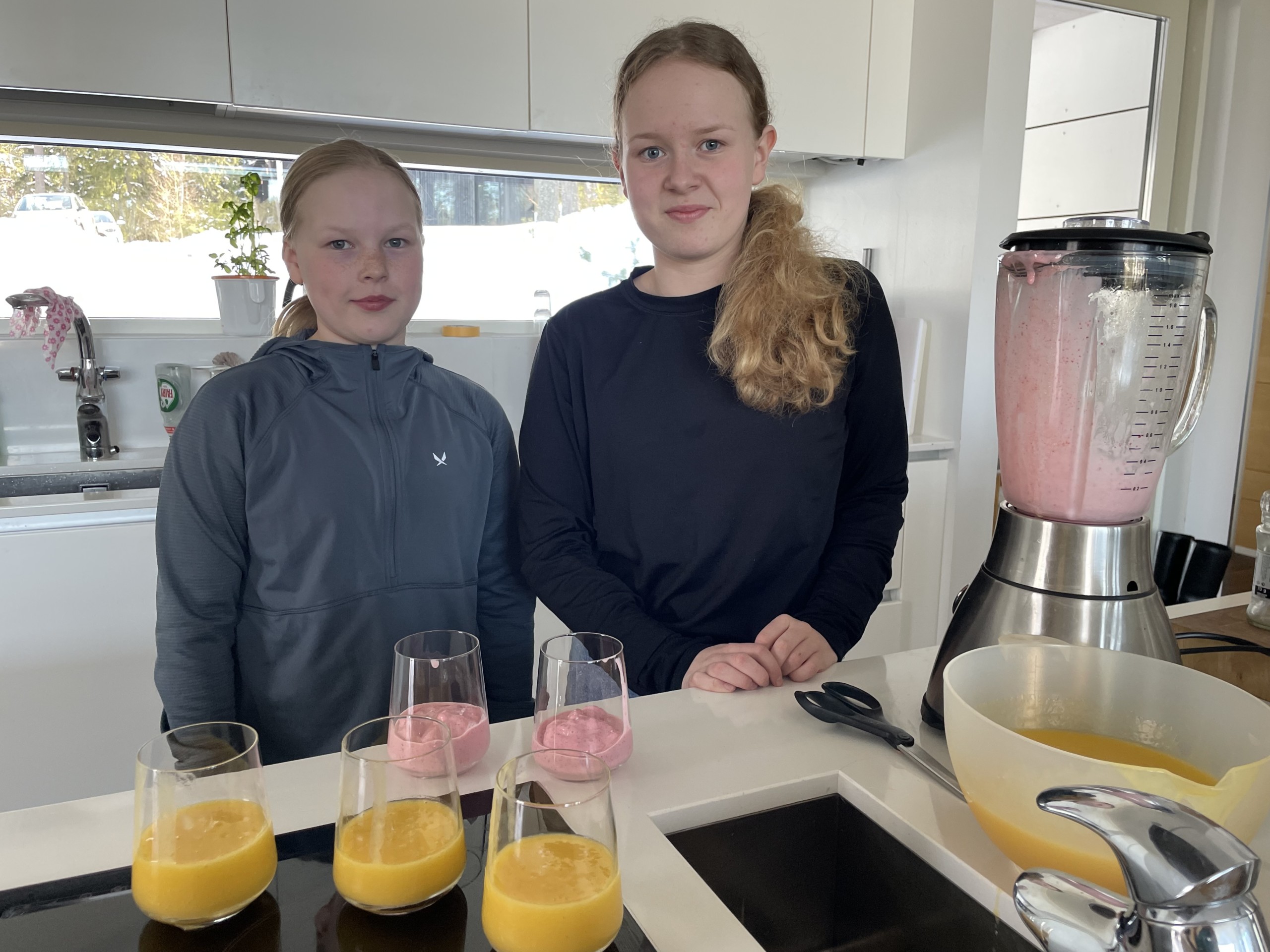 två flickor i ett kök. Flera glas med smoothie på bordet och en mixer.