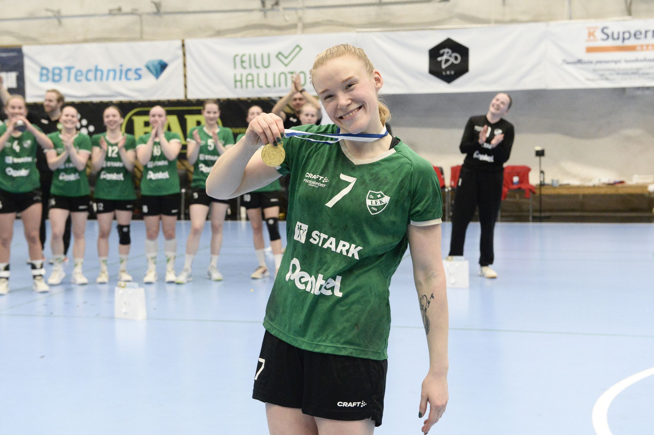 en leende kvinna i grön tröja visar upp en guldmedalj
