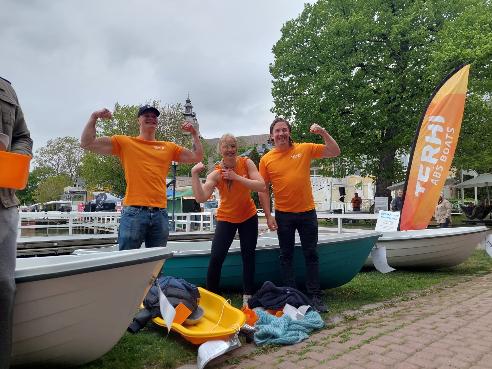 Tre personer i orange tröjor poserar framför en roddbåt