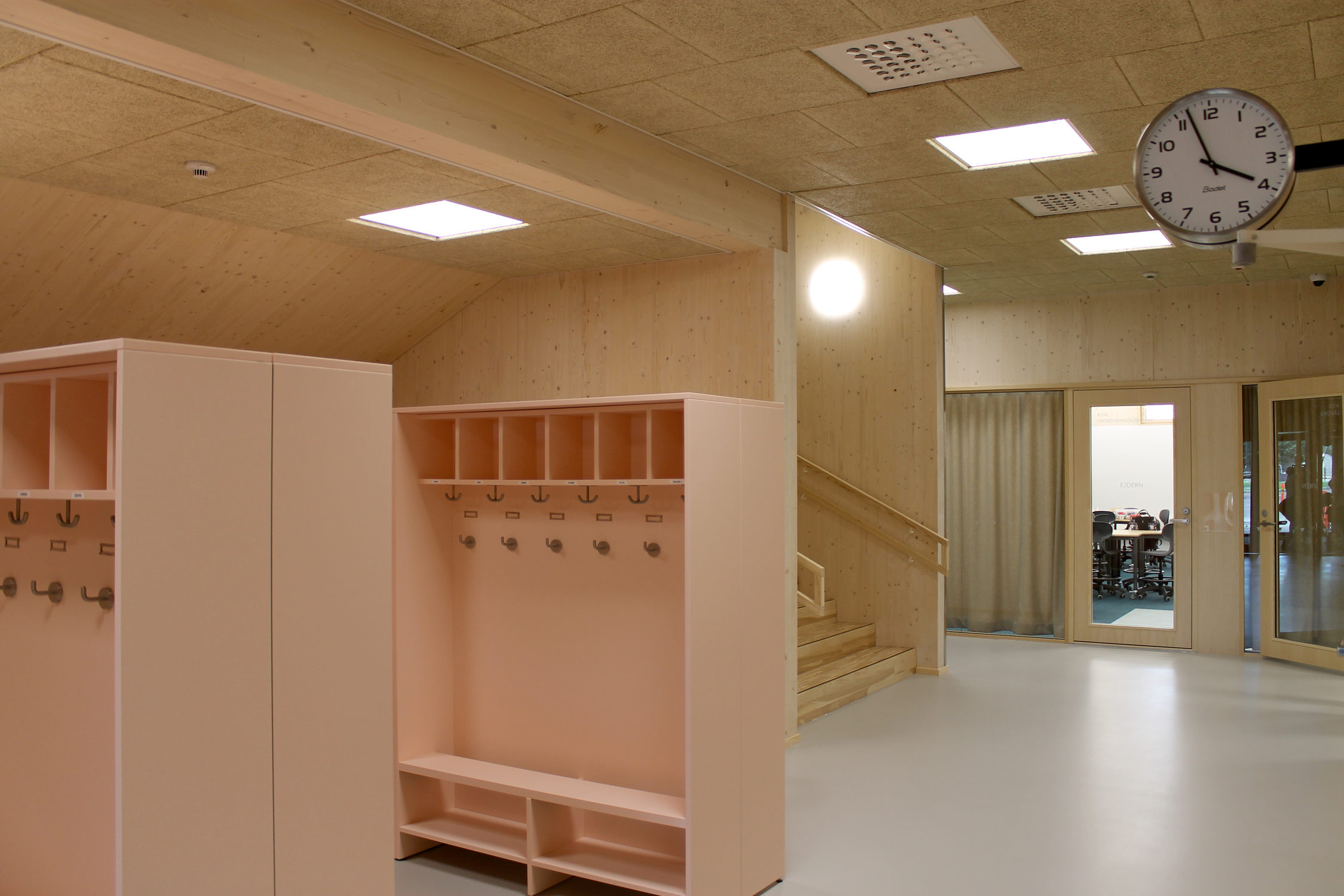 En skolas entréhall med öppna skåp för ytterkläder, skåpen är ljusrosa. Väggarna i skolan är av ljust trä.