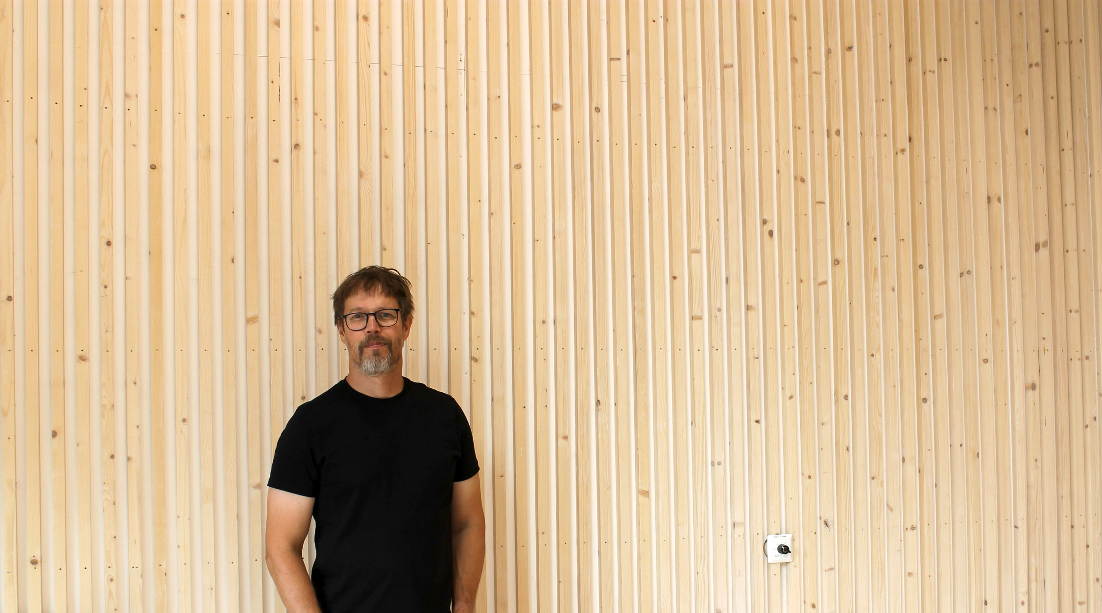 En man står framför en vägg i ljust trä, mannen är arkitekt till skolbyggnaden han står i.