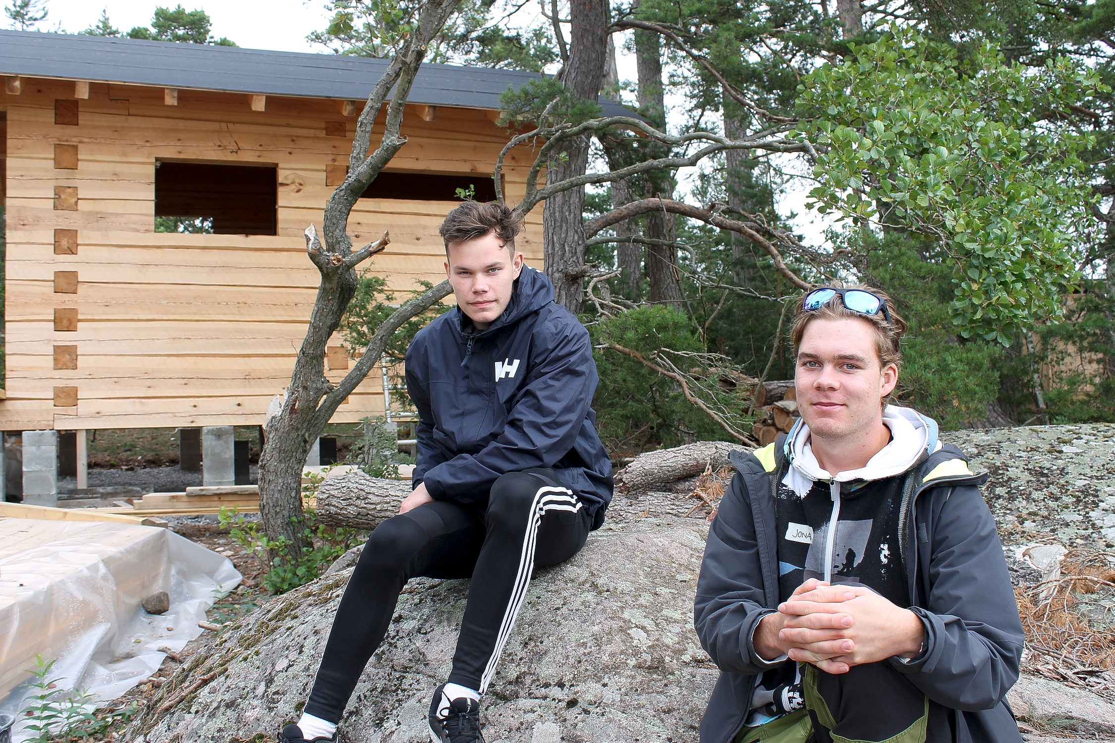 Två pojkar sitter bredvid varandra på en klippa i Bromarvs skärgård. De är studernade vid Novia i Ekenäs och är i dag på utflykt med en intensivkurs till några stockstugor som S-trä bygger. En av stugorna syns i bakgrunden på bilden.