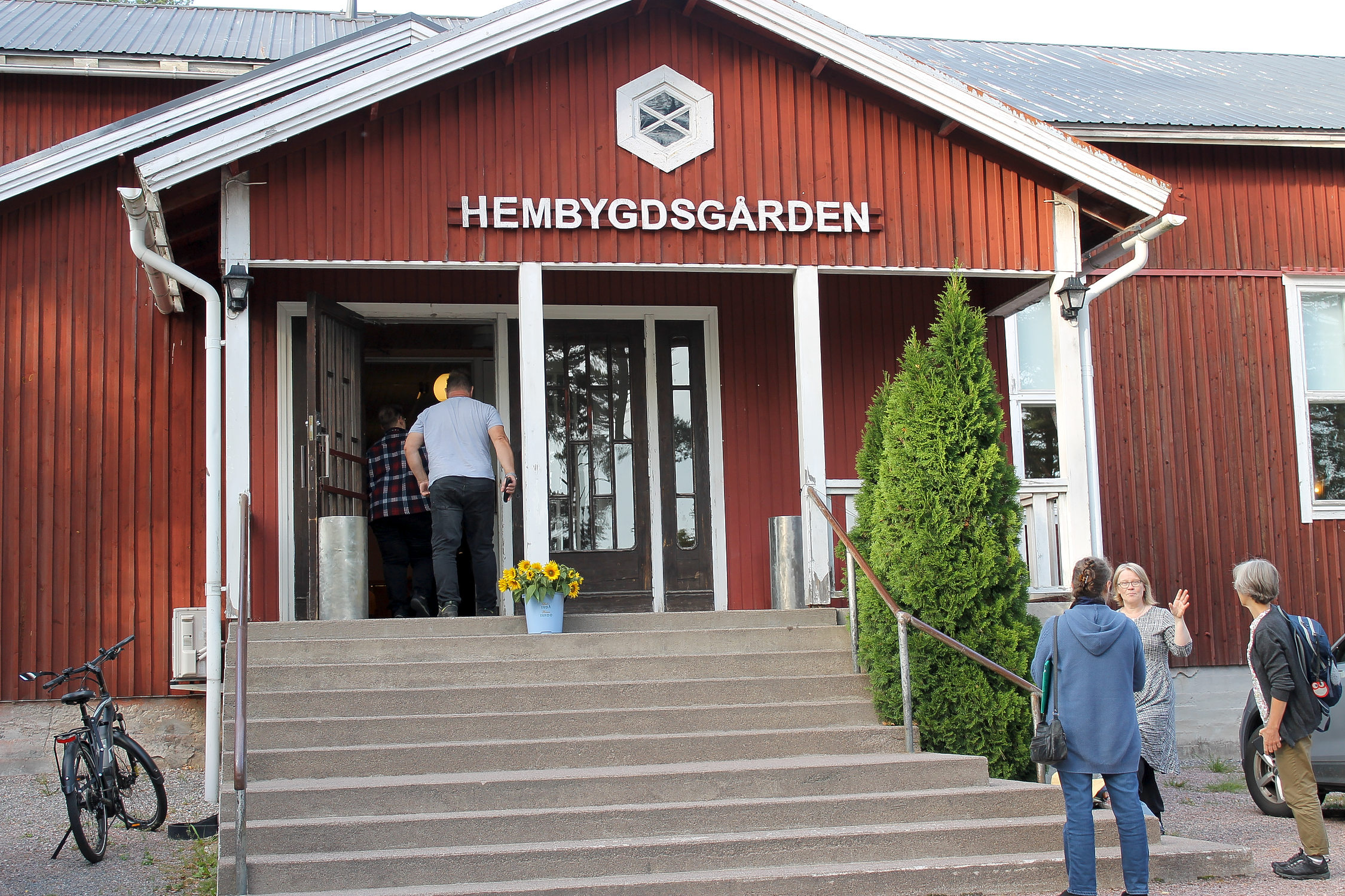 Hembygdsgården i Ingå. Ett rött hus med skylten "Hembygdsgård" över ingången. Några personer går in i huset och några står vid trappan till huset och diskuterar.