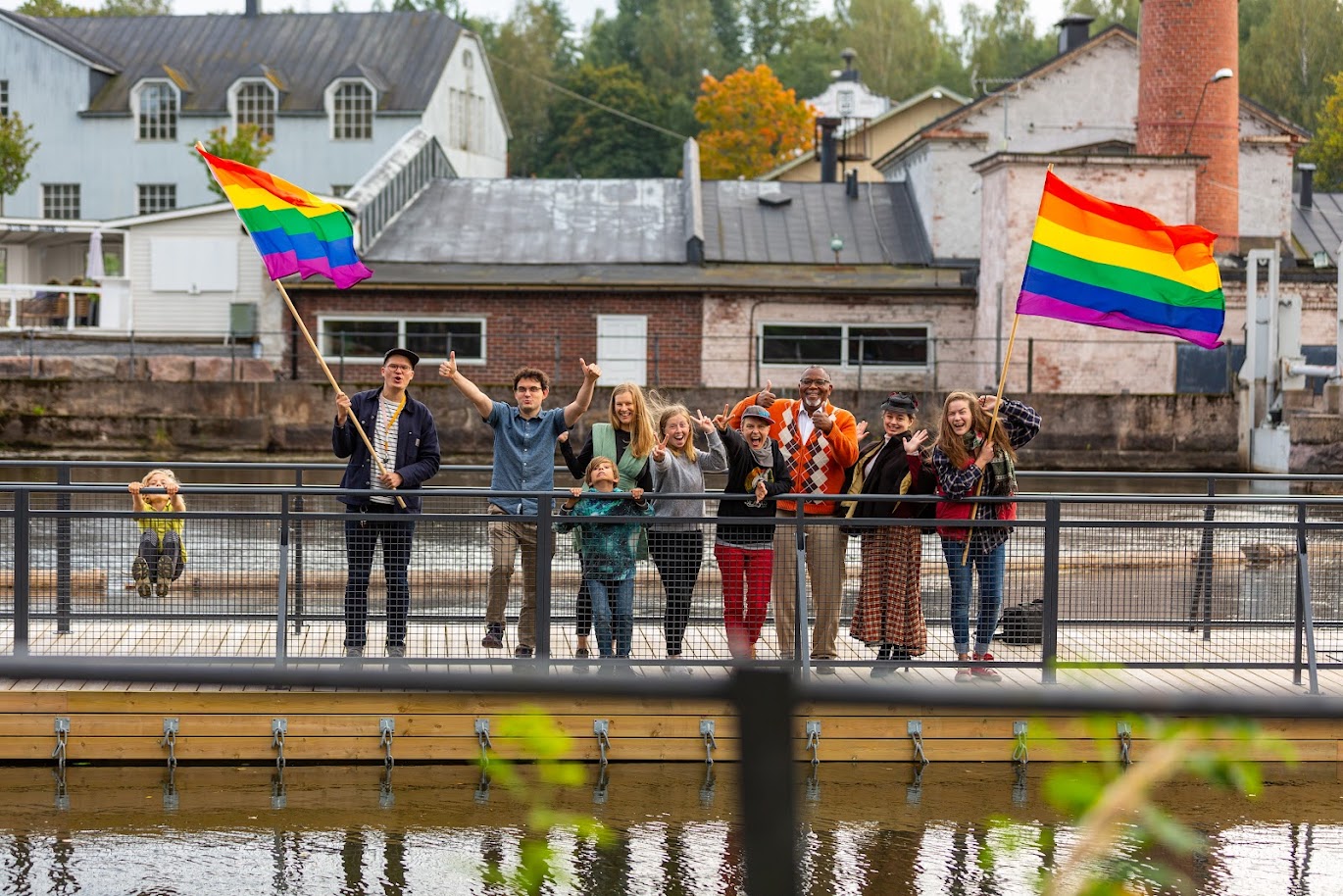 En grupp människor står på en bro och viftar med regnbågsflaggor och ser glada ut.