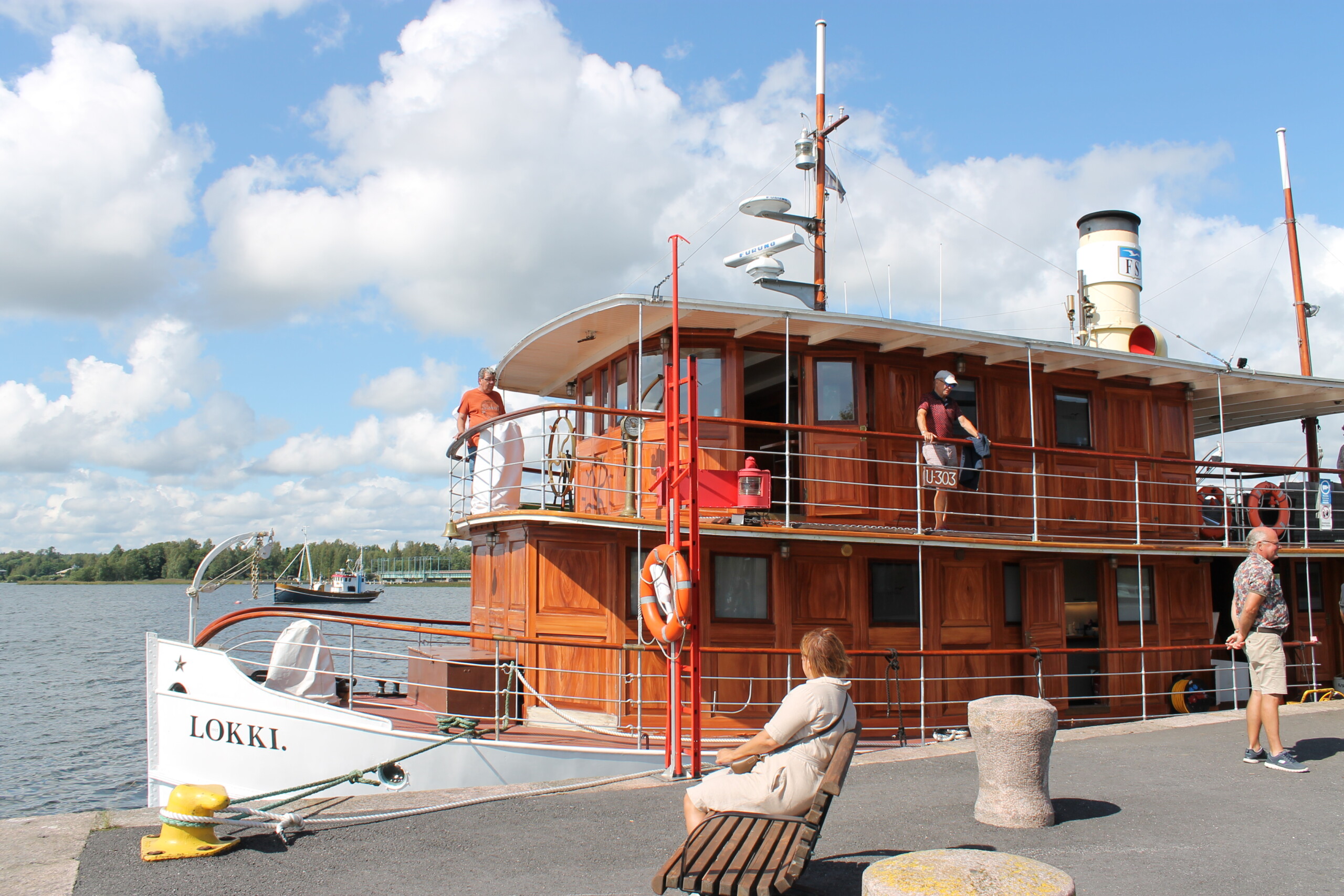 En gammal ångbåt står i en hamn. Vatten och himmel syns i bakgrunden. På båten går flera människor omkring. På en bänk vid båten sitter en kvinna på en bänk.