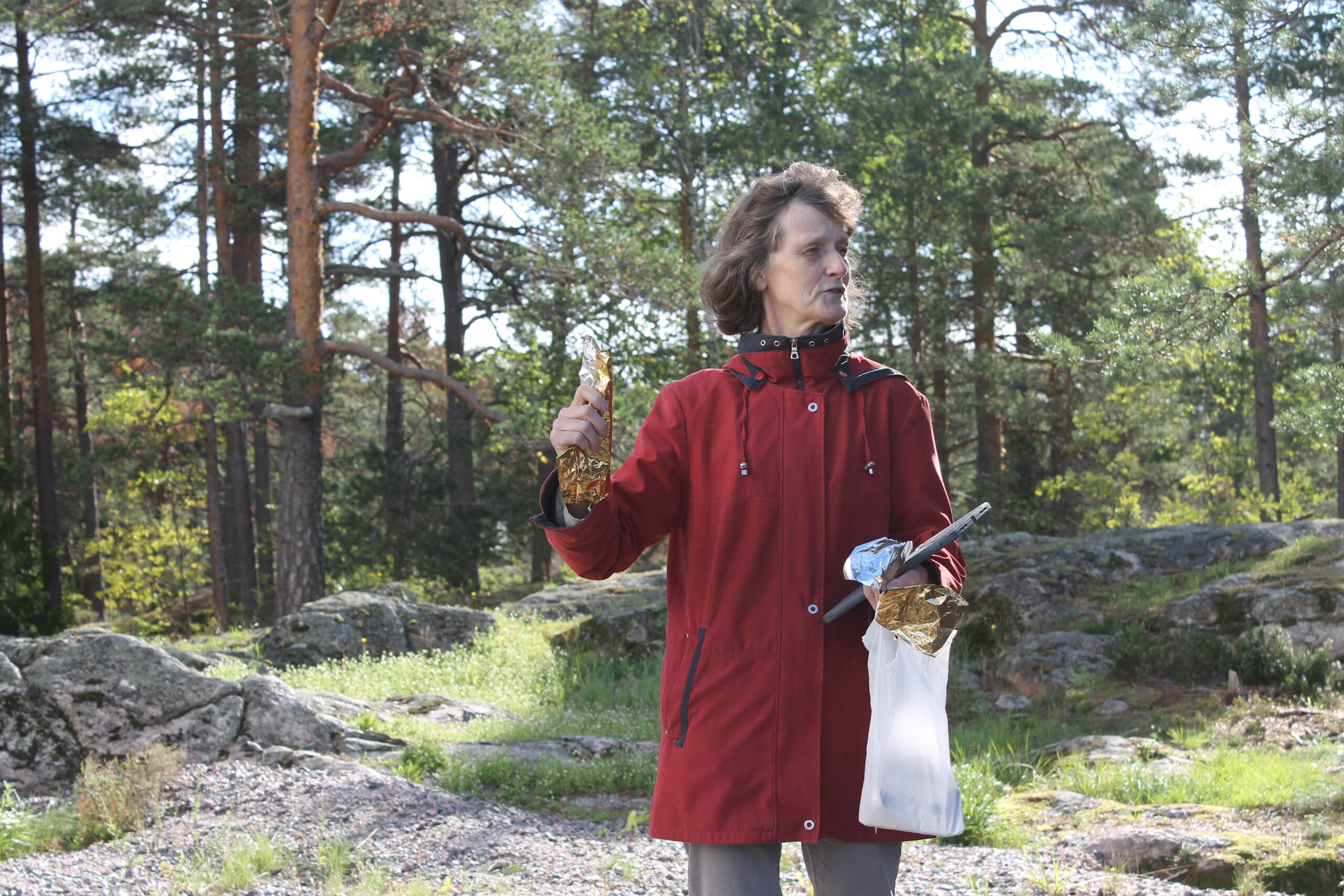 En kvinna står utomhus med skog i bakgrunden. Hon håller i blänkande papper och pratar inför en grupp elever som ej syns på bilden.
