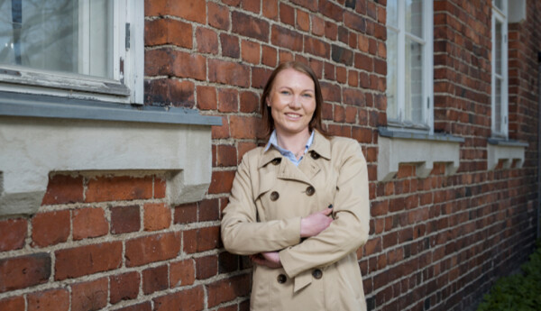 Kvinna i beige trenchcoat står med armarna i kors och lutar mot en röd tegelvägg.