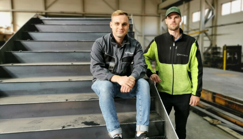 Tvä män i verkstadshall och den ena mannen sitter på en svetsad trappa.