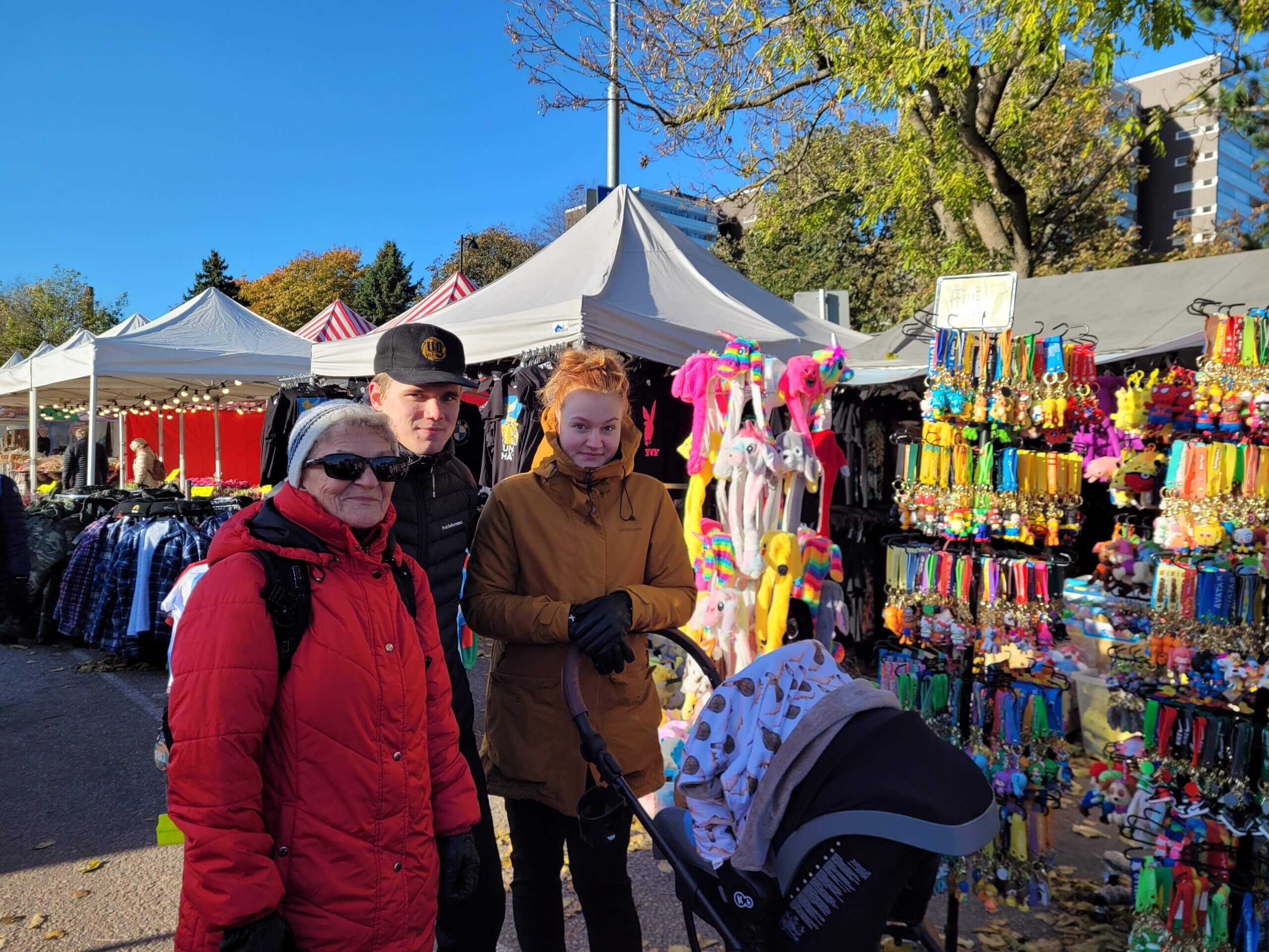 En äldre kvinna samt ett ungt par med en barnvagn ler mot kameran i solskenet på marknaden.