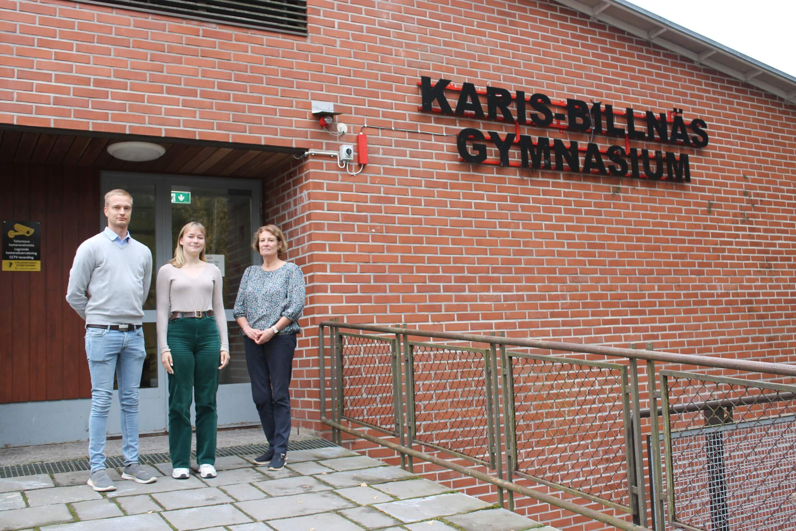 En man, en ung kvinna och en kvinna står bredvid varandra utanför en röd tegelbyggnad med skylten "Karis-Billnäs Gymnasium". De arbetar alla på skolans skoltidning.