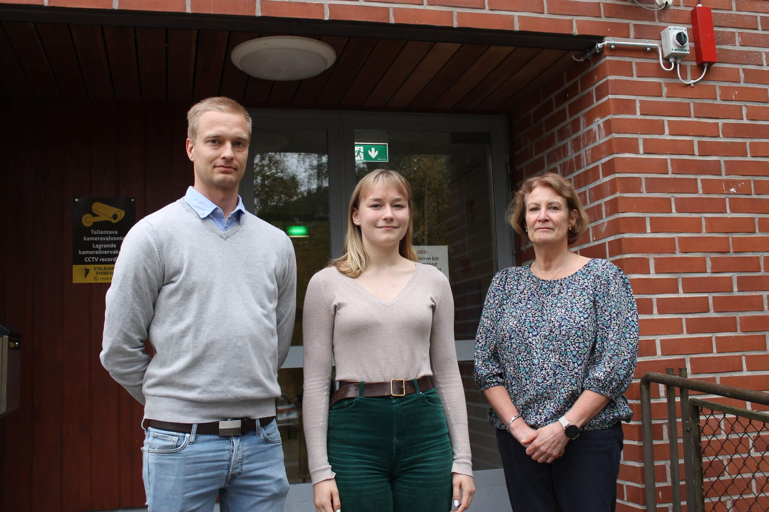 En man , en ung kvinna och en kvinna står bredvid varandra utanför en röd tegelbyggnad som är Karis-Billnäs gymnasium. Mannen och kvinnan är lärare på skolan och den unga kvinnan är studerande. Alla tre arbetar på skolans skoltidning.