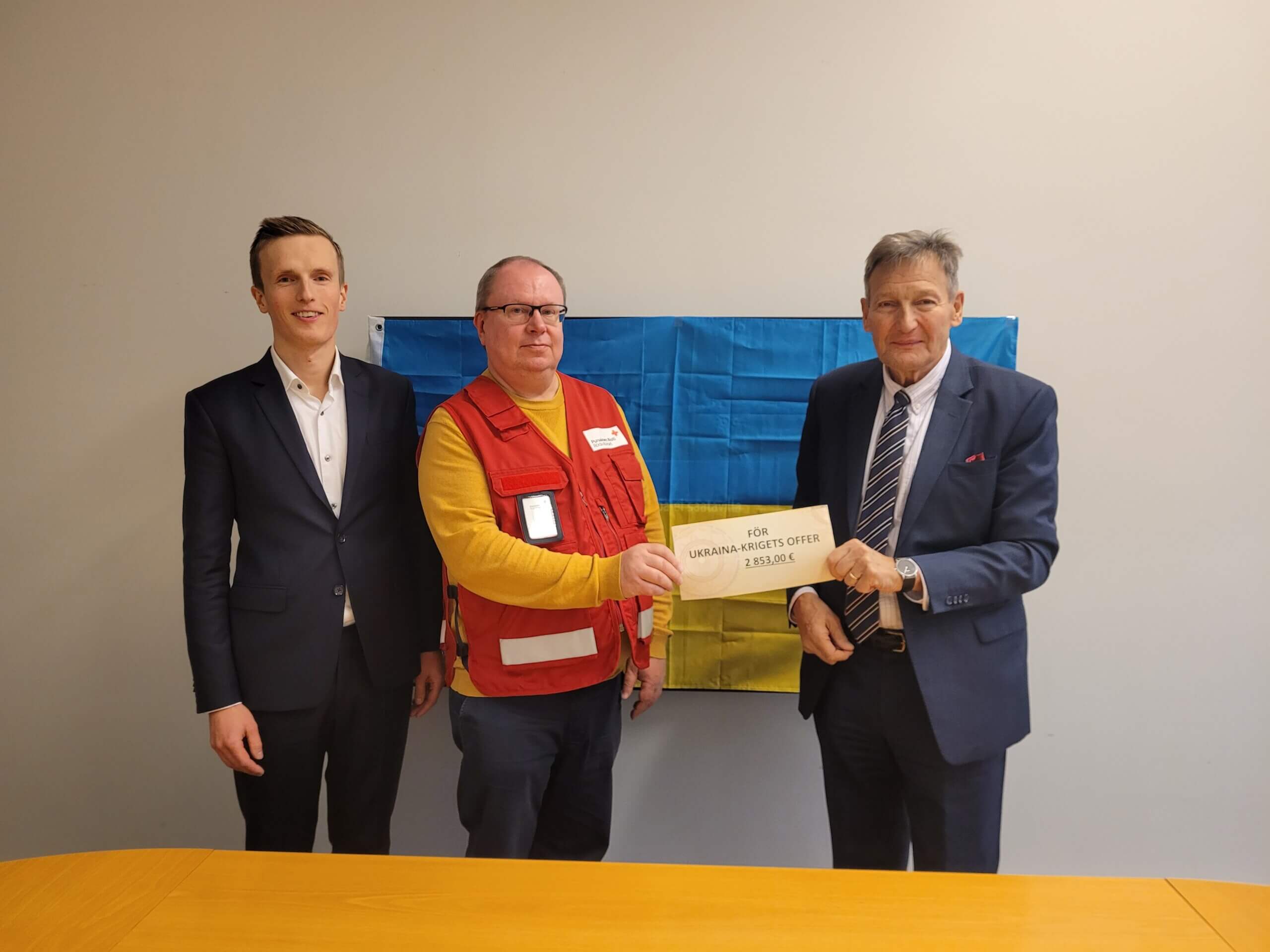 Tre män står framför en Ukrainaflagga som är upphängd på väggen. I handen håller de i en check med donationssumman till Ukrainas krigsoffer.