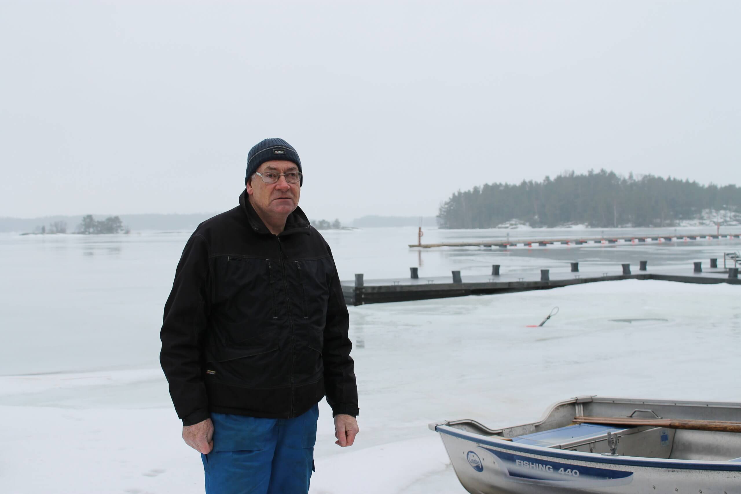En äldre man står utomhus vid hamnen Baggö i Ekenäs skärgård. Det är snö på marken och i bakgrunden ser man en ö, några bryggor, dimma och fruset vatten.