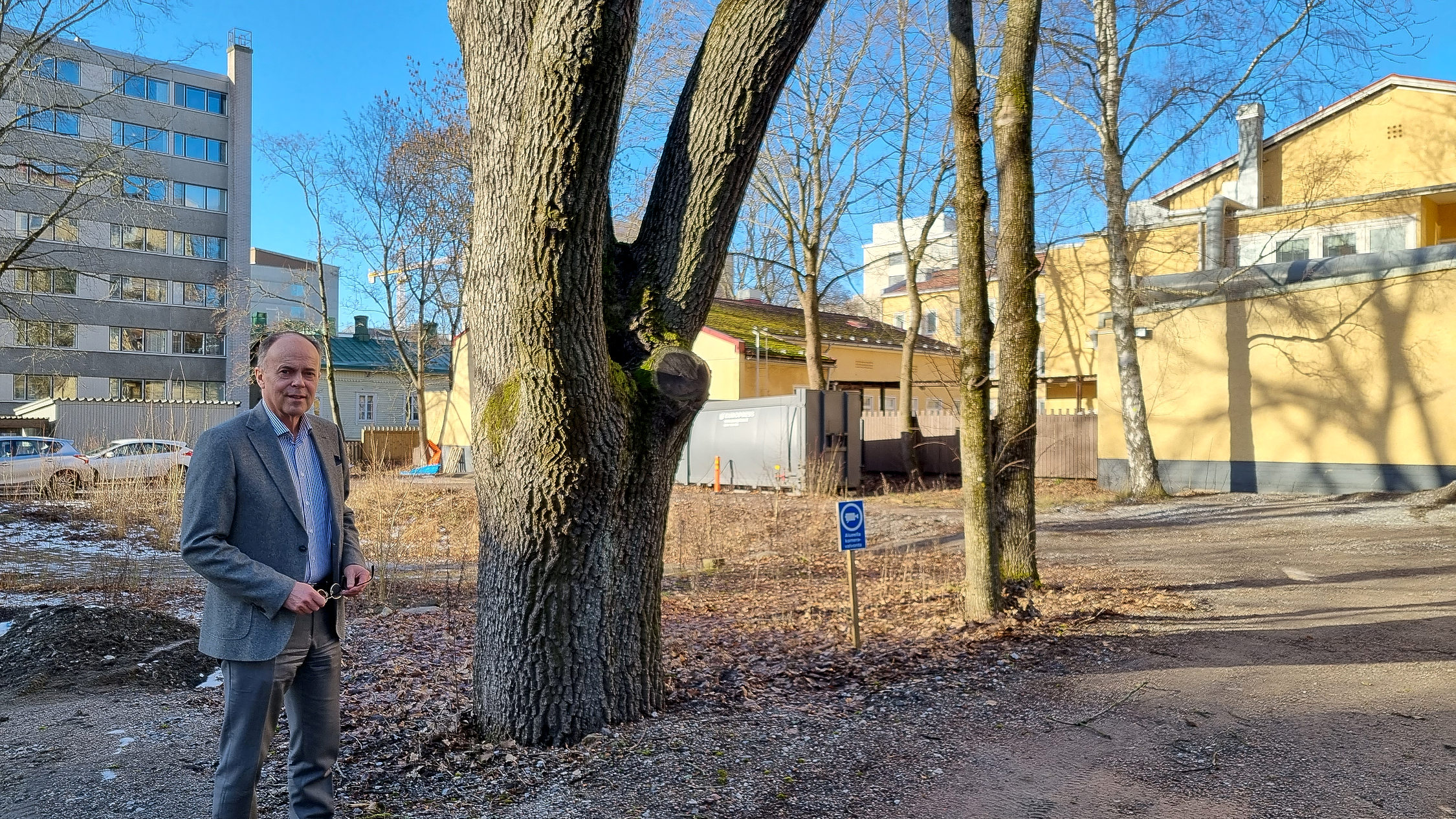man står på innergård i vårsol med stort träd bredvid sig