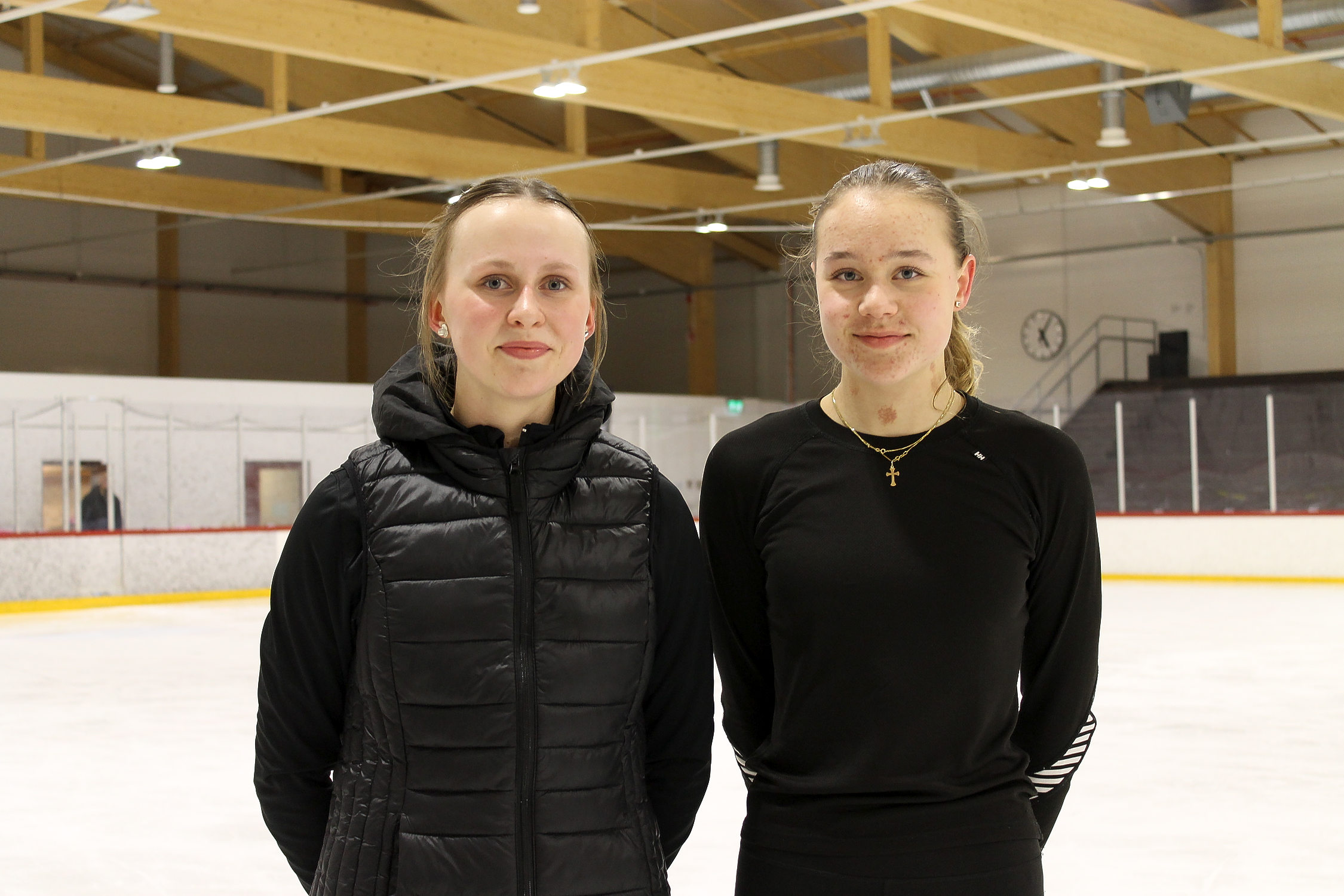 Två unga flickor klädda i svarta träningskläder står på isen i Karis ishall. Flickorna tittar in i kameran och ler. De är båda elever i Hurja Piruettis kurs i konståkning.