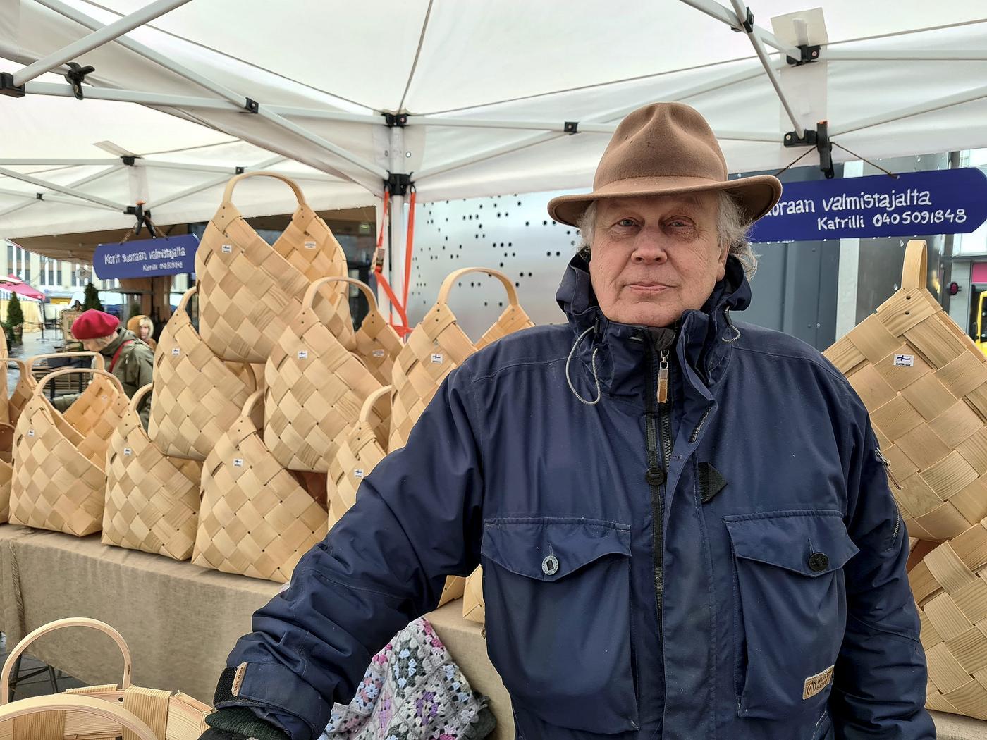 En äldre man i ytterrock och hatt står bland handflätade korgar