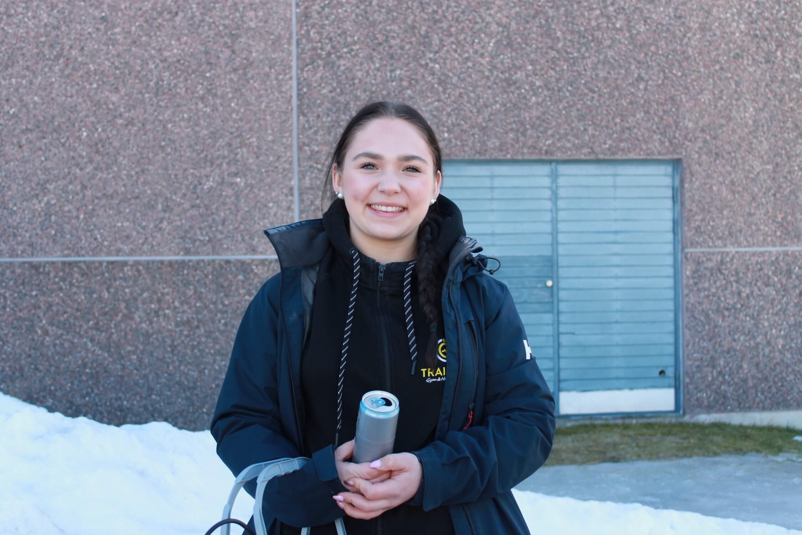 En ung kvinna står och tittar in i kameran och ler. Kvinnan står utomhus och i bakgrunden ser man en vägg av en byggnad. Kvinnan har nyss skrivit vårens första studentskrivning i Ekenäs.