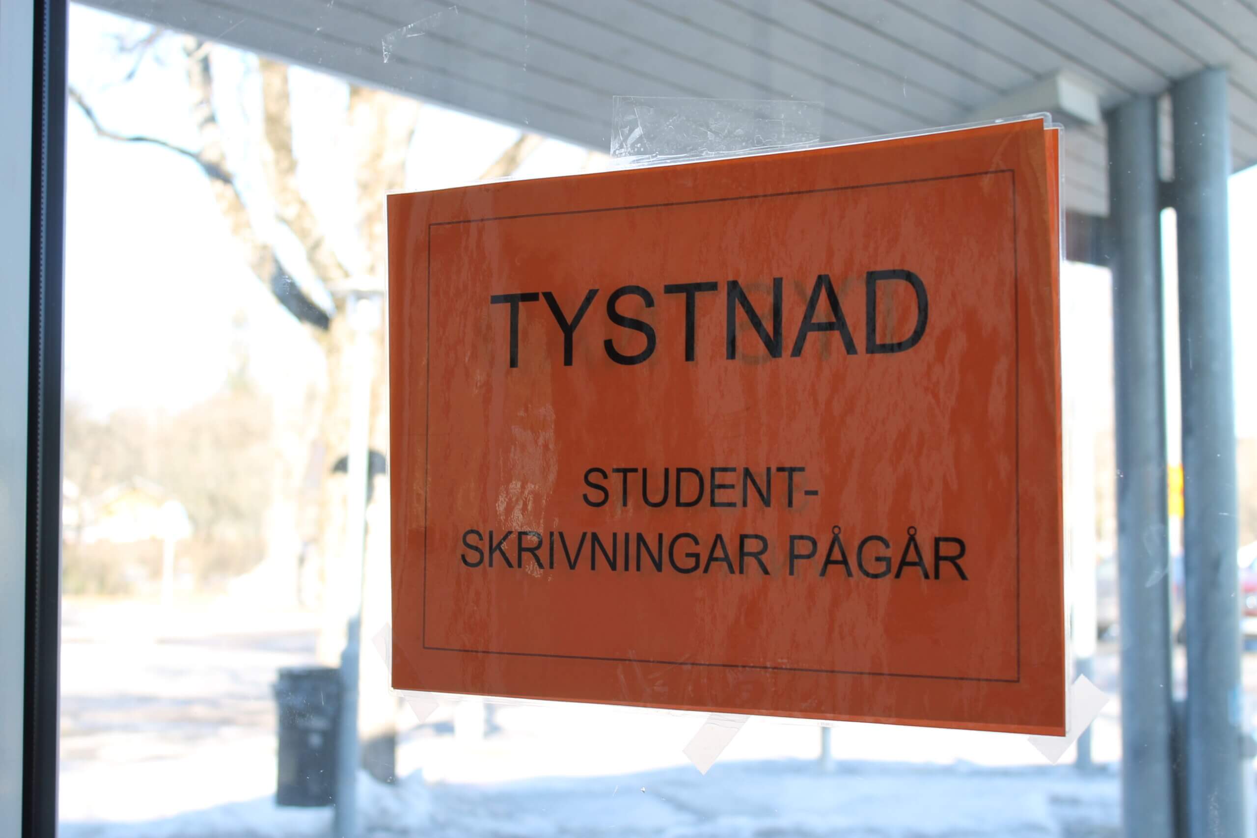 Närbild på en skylt som är fast på ett fönster. På skylten står det "Tystnad. Studentskrivningar pågår." Skylten är orange.