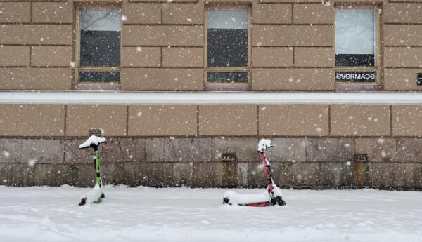 Två elsparkcyklar står parkerade framför en husfasad mitt i snöyra.