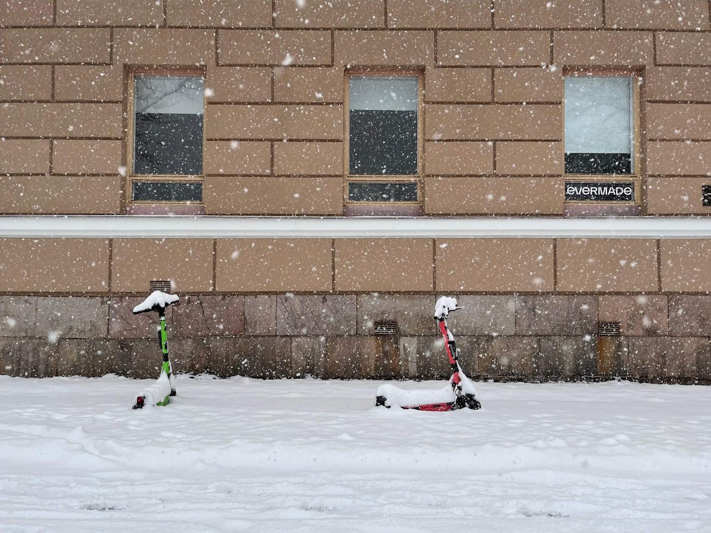 Två elsparkcyklar står parkerade framför en husfasad mitt i snöyra.