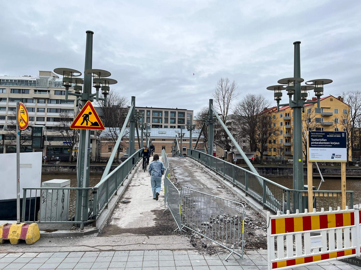 Gångbro med staket som löper i mitten. På vänster sida om bron är en fotgängare och en cyklist, på höger sida en uppskrapad vägyta