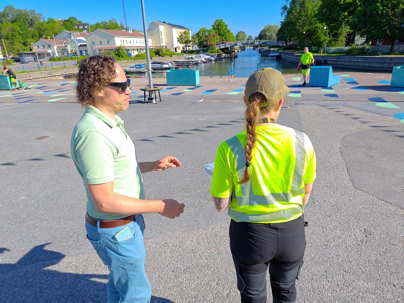 Man med sida mot kameran och kvinna i gul väst med ryggen mot kameran som tittar ut över torget.