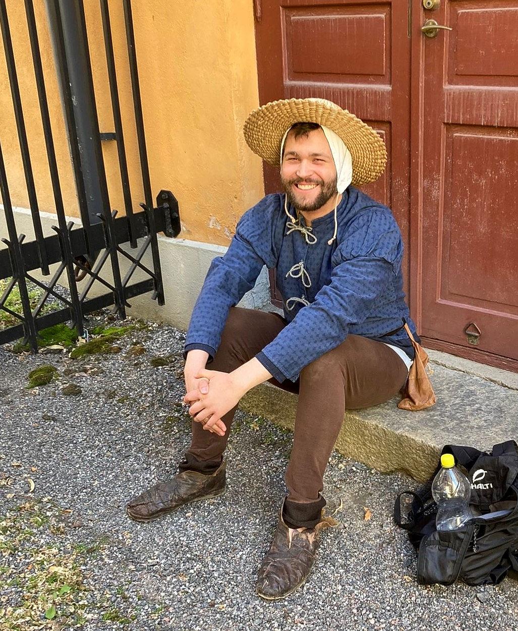 Tapio Helminen i sin kostym för pjäsen. Han har på sig bruna byxor, en blå skjorta och en halmhatt. 