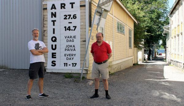 Två män står bredvid varandra och tittar in i kameran. Männen står utomhus och solen lyser på gatan bakom männen. Bakom dem finns en grusväg och ett stort gult trähus med en skylt som det står "Art Antiques" på.