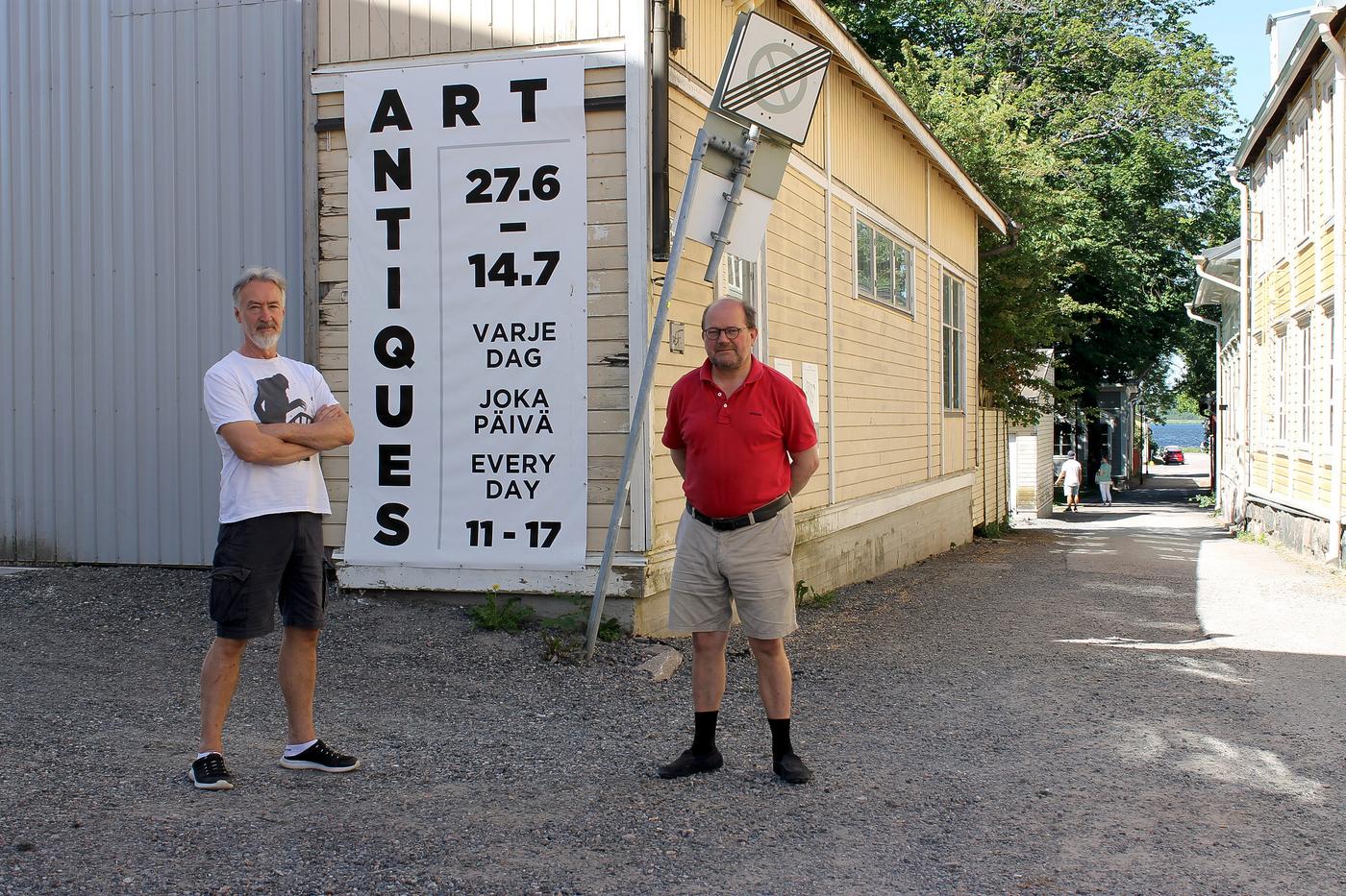 Två män står bredvid varandra och tittar in i kameran. Männen står utomhus och solen lyser på gatan bakom männen. Bakom dem finns en grusväg och ett stort gult trähus med en skylt som det står "Art Antiques" på.