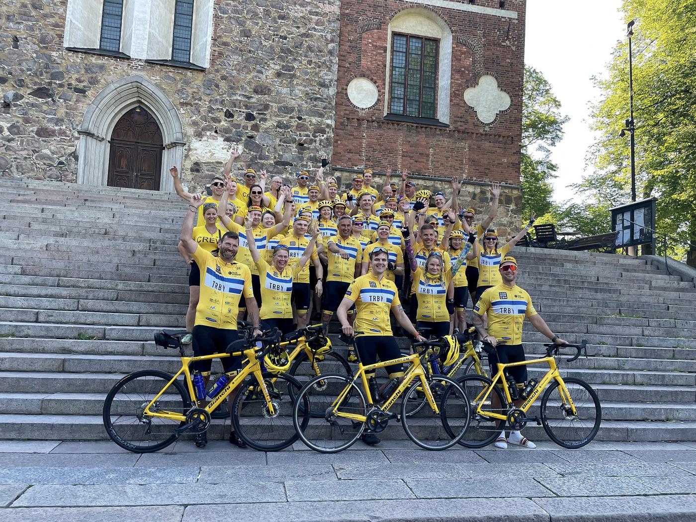 Bild på teamet från åbo framför domkyrkan före start. Alla har gula lagskjortor och cyklar.