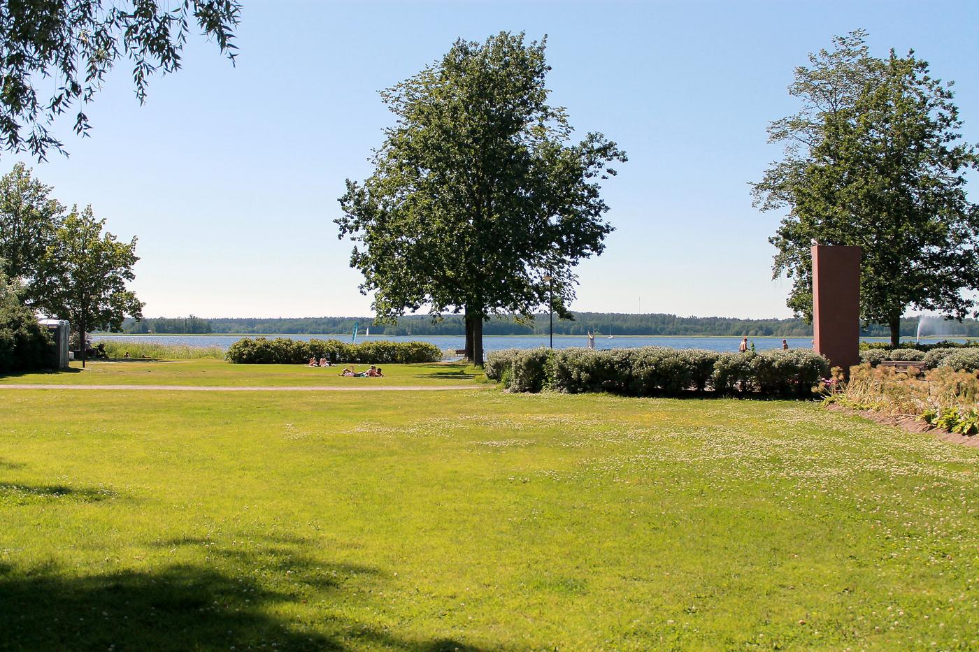 En grön gräsmatta och några gröna träd. I bakgrunden ser man havet och några människor som ligger nära vattnet och solar.