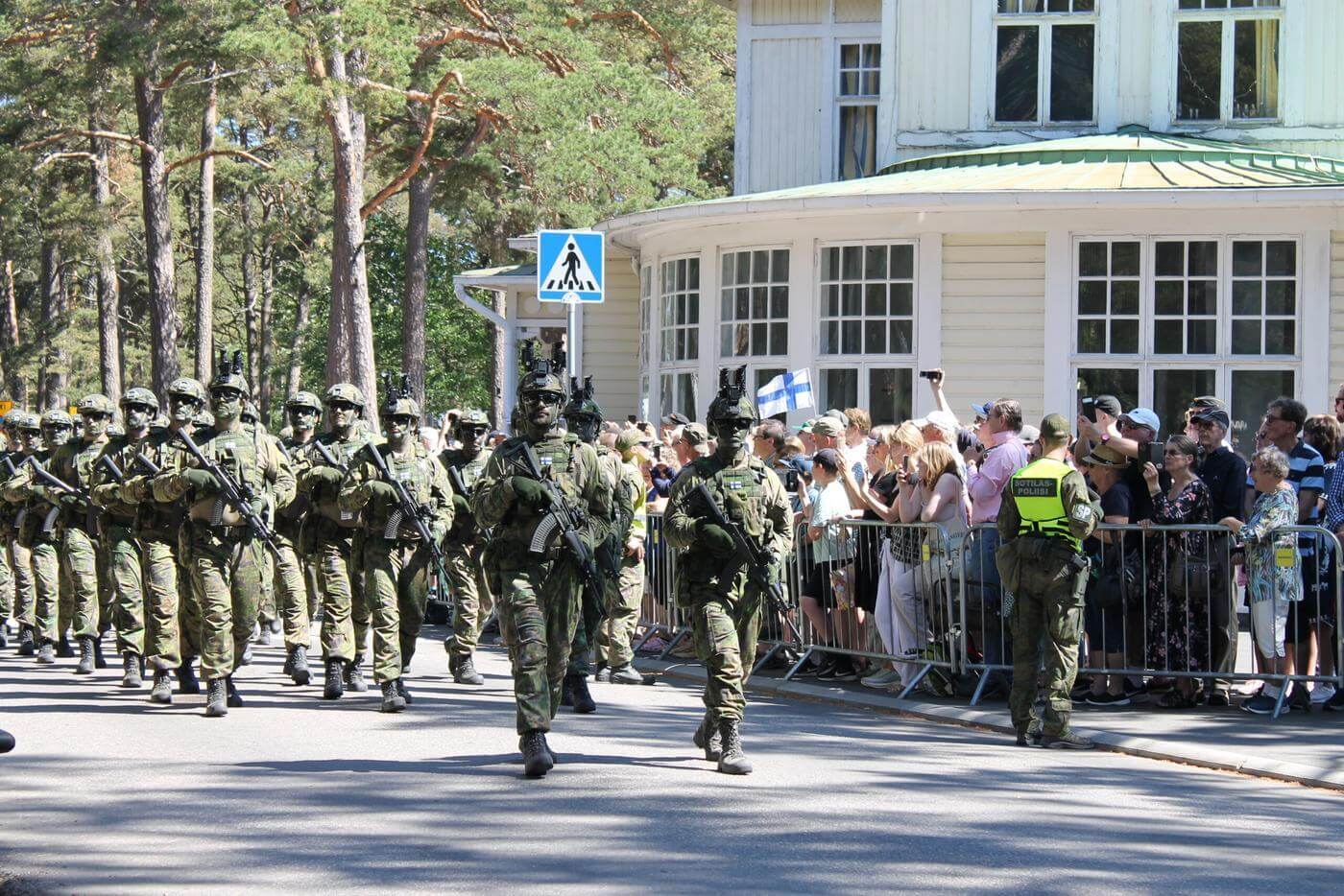 Militärklädd trupp marscherar förbi Casino i Hangö. I bakgrunden ser man människor som samlats för att se paraden.