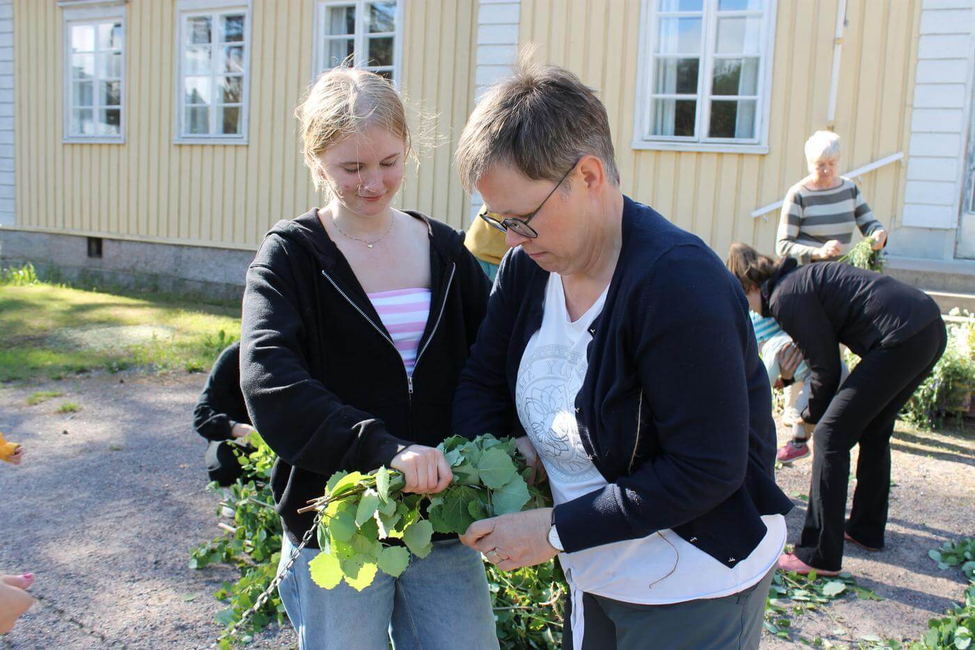 Två kvinnor står utomhus och sköter lövningen till midsommarstången i Tenala.