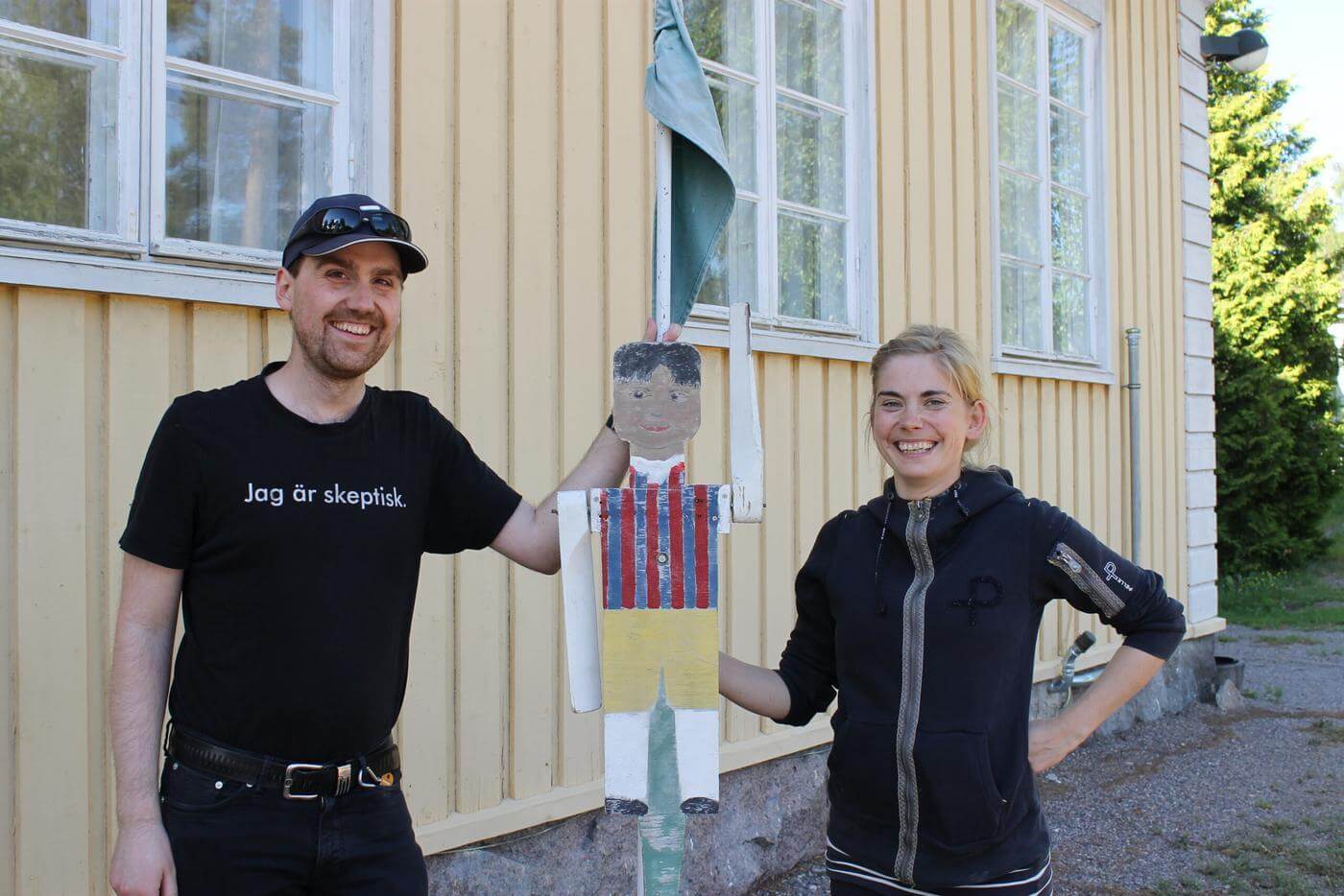 En man och en kvinna står utomhus. Mellan sig håller de upp en gubbe klädd i Tenala folkdräkt. Gubben är gjord av trä och ska vara på toppen av midsommarstången i byn.