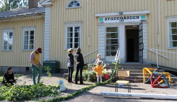 Ett stort gult trähus med skylten Bygdegården ovanför dörren. Utanför huset står människor och sköter lövningen och bindningen av midsommarstången i Tenala.